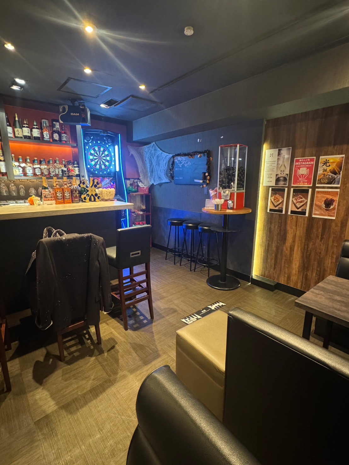 あたしンちの店内。オレンジに照らされたボトル棚のバーカウンター、青く光るダーツマシン、スツール付きのハイテーブル、黒いレザーのソファ。木目の壁に「あたしンち INSTAGRAM」のポスター。